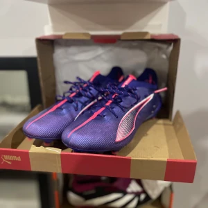 Puma ultra ultimate  - Snygga lila fotbollsskor från Puma med rosa detaljer och snörning. Skorna har en modern design med en slitstark sula och är perfekta för fotbollsplanen. De har en strömlinjeformad form för optimal prestanda.