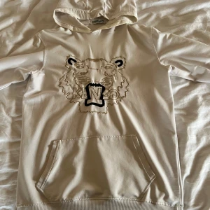 Vit hoodie från Kenzo med tigerbroderi - Säljer en stilren vit hoodie från Kenzo med ett coolt tigerbroderi på framsidan. Hoodien har en praktisk magficka och en bekväm huva. Perfekt för en avslappnad look.