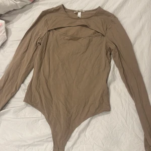 Beige långärmad bodysuit - Säljer en stilren beige långärmad bodysuit med unik design. Den har en intressant detalj vid halsen som ger en modern touch. Perfekt för att kombinera med olika outfits.