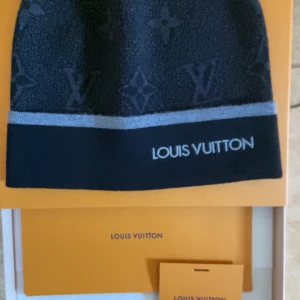 Svart mössa från Louis Vuitton Kvitto Finns! - Elegant svart mössa från Louis Vuitton med grå detaljer och det ikoniska LV-mönstret. Mössan har en broderad logotyp och är tillverkad i en mjuk ullblandning. Perfekt för att hålla stilen under kallare dagar.