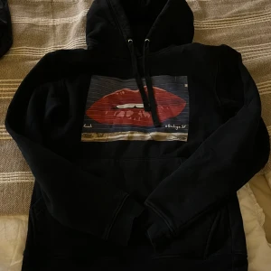 Svart hoodie från The Cool Elephant - Säljer en svart hoodie från The Cool Elephant med ett tryck av röda läppar på framsidan. Hoodien har en klassisk design med dragsko i huvan och en stor ficka framtill. Perfekt för en avslappnad stil.