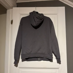 Grå hoodie med dragkedja - Säljer en grå hoodie med dragkedja och textdetalj på bröstet. Tröjan har en justerbar huva och fickor framtill. Perfekt för en avslappnad stil.