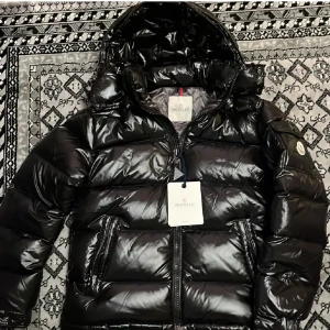 Svart pufferjacka från Moncler - Snygg svart pufferjacka från Moncler med glansig finish och huva. Jackan har dragkedja framtill och Moncler-logga på ärmen. Perfekt för kyliga dagar.