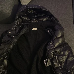 Svart dunjacka med huva - Säljer en snygg svart dunjacka från Moncler med huva. Jackan har en quiltad design och dragkedja framtill. Perfekt för kyliga dagar och ger en stilren look.