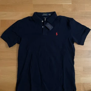 Mörkblå pikétröja från Ralph Lauren - Snygg mörkblå pikétröja från Ralph Lauren med klassisk krage och knappar. Den har en röd broderad logga på bröstet. Perfekt för en stilren look.