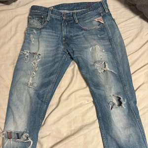 Blå jeans från Replay - Säljer ett par blå Replay Anbass jeans med slitna detaljer och en cool, distressed look. De har en klassisk femficksdesign och knappgylf. Perfekta för en avslappnad stil.