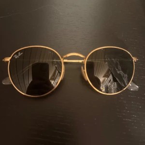 Runda solglasögon från Ray-Ban - Snygga runda solglasögon från Ray-Ban med guldfärgad metallram och mörka glas. Perfekta för en stilren look. De har justerbara nässadlar för extra komfort. Nypris 1750kr