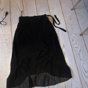 Svart omlottkjol - Snygg svart omlottkjol med knytband i midjan. Kjolen har en elegant volangdetalj och är perfekt för en stilren look. Den är i en längre modell och passar bra till olika tillfällen.