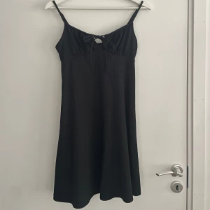 Svart klänning från Gina Tricot - Snygg svart klänning från Gina Tricot med smala axelband och knytning framtill. Perfekt för en stilren look. Klänningen har en enkel och elegant design som passar till många tillfällen.