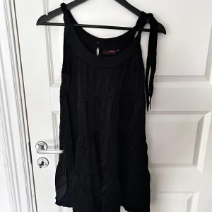 Svart klänning från Zoul - Snygg svart klänning från Zoul med axelbandslös design och volangdetaljer. Perfekt för en stilren look. Klänningen har en luftig passform och är tillverkad i ett mjukt material.