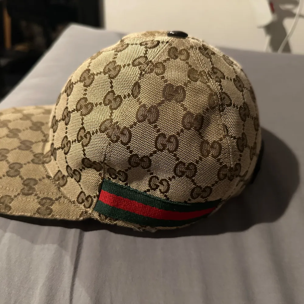 Snygg beige keps från Gucci med det ikoniska monogrammönstret. Kepsen har en justerbar rem i skinn baktill och en grön och röd randdetalj på sidan. Perfekt för att ge din outfit en lyxig touch.. Asusteet.