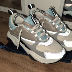 Grå och beige sneakers från Dior - Snygga sneakers från Dior i en kombination av grått, beige och vitt med blå detaljer. Skorna har en chunky sula och snörning framtill. Perfekta för en stilren look. Skorna är stora i storleken den passar 41-42. Priser går att sänkas vid snabb affär 