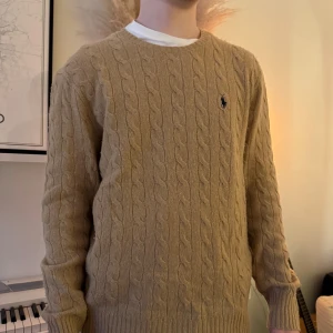 Beige kabelstickad tröja från Ralph Lauren - Säljer en stilren beige kabelstickad tröja från Ralph Lauren. Tröjan har en klassisk rund halsringning och är prydd med den ikoniska logotypen på bröstet. Perfekt för en elegant och avslappnad look.