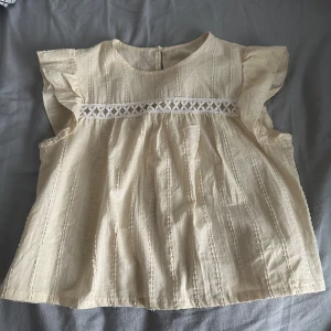 Beige blus med spetsdetalj - Charmig beige blus med korta volangärmar och vacker spetsdetalj över bröstet. Blusen har en rund halsringning och ett diskret mönster i tyget som ger en fin struktur. Perfekt för en somrig look!