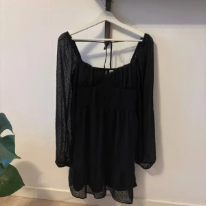 Svart klänning med prickigt mönster - Snygg svart klänning med långa ärmar och prickigt mönster. Klänningen har en volangdetalj vid fållen och en knytning i ryggen. Perfekt för en stilren look.