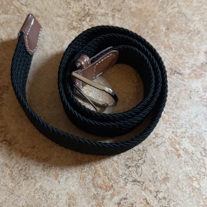 Svart flätat bälte med bruna detaljer - Snyggt svart flätat bälte med bruna läderdetaljer och silverfärgat spänne. Perfekt för att ge en stilren touch till din outfit. Bältet har en klassisk design med kontrasterande sömmar på läderdetaljerna.