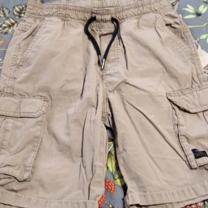 Beige cargo shorts från Kappahl - Säljer ett par beige cargo shorts från Kappahl med elastisk midja och snörning. Shortsen har flera praktiska fickor med lock på sidorna. Storlek 152.