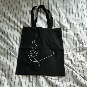 Svart shoppingväska  - Snygg svart shoppingväska med ett stilrent vitt ansiktsmönster. Perfekt för att bära med sig det nödvändigaste. Väskan har två långa handtag för enkel bärning 💕