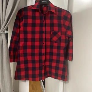 Röd och svart rutig skjorta från Gina Tricot - Snygg röd och svart rutig skjorta från Gina Tricot med knappar framtill och en bröstficka. Perfekt för en avslappnad stil. Skjortan har långa ärmar och en klassisk krage.