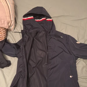 Mörkblå jacka med huva från Moncler - Säljer en snygg mörkblå jacka från Moncler med huva och dragkedja. Jackan har en stilren design med Moncler-logga på ärmen och röd-vit randig detalj på huvan. Perfekt för våren. NFC funkar och tar dig direkt till varan på hemsidan 