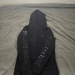 Svart hoodie från Icono - Säljer en svart hoodie från Icono med texten 'ICONO COUTURE' längs båda armarna. Hoodien har en dragkedja och en snygg logotyp på bröstet. 