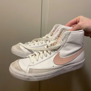 Vita Nike sneakers med rosa detaljer - Snygga vita Nike sneakers med rosa swoosh och detaljer. Skorna har hög modell med snörning och en klassisk design. Perfekta för en stilren look. Nypris ~ 900/1000kr