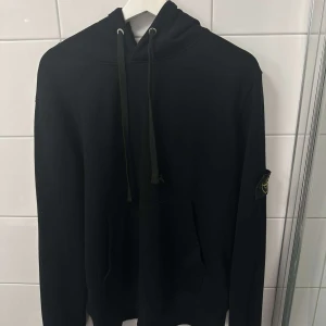 Svart hoodie med logga på ärmen - Snygg svart hoodie med justerbar dragsko i huvan och en logga på vänster ärm. Perfekt för en avslappnad stil. Den har en klassisk känguruficka framtill.
