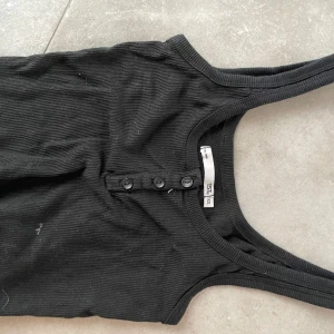 Svart ribbad linnetopp - Säljer en svart ribbad linnetopp med knappar framtill. Toppen är croppad och har breda axelband, perfekt för en stilren look. Passar bra till både jeans och kjol.