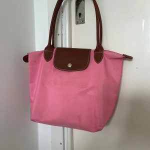 Äkta rosa Longchamp - Snygg äkta rosa Longchamp med bruna handtag och detaljer. Väskan har en praktisk dragkedja och en knappstängning framtill. Perfekt för att bära med sig det nödvändigaste med stil. Den har en guldig dragkedjedetalj som ger en extra touch av elegans. Finns lite fläckar på den.