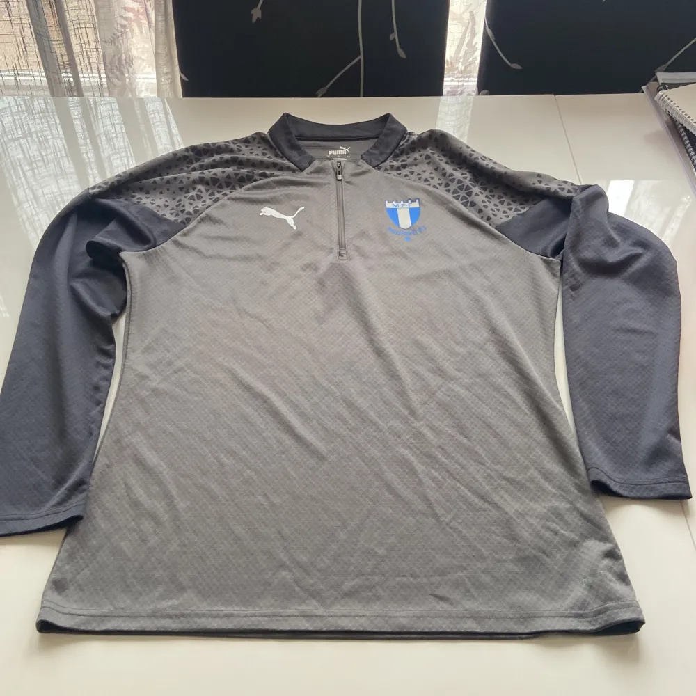Säljer en grå långärmad träningströja från Puma med Malmö FF-logga på bröstet. Tröjan har ett diskret mönster på axlarna och en halv dragkedja framtill. Perfekt för fotbollsträning eller andra sportaktiviteter.. Neuletakit & Villapaidat.
