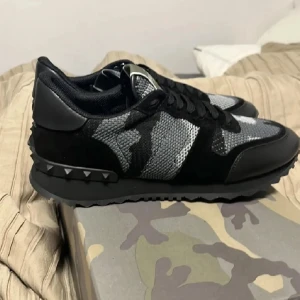 Valentino Garavani sneakers i svart och grått - Snygga Valentino Garavani sneakers i svart och grått med ett camouflage-mönster. Strlk 43 (passar 42). Skick 8/10. Kan mötas upp i Stockholm! Box och dustbag medföljer!