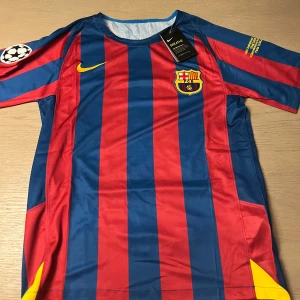 Blå och röd fotbollströja från Nike - Säljer en klassisk FC Barcelona fotbollströja från Nike med Ronaldinho och nummer 10 på ryggen. Tröjan har korta ärmar och är randig i blått och rött med klubbens emblem på bröstet och Champions League-märke på ärmen.