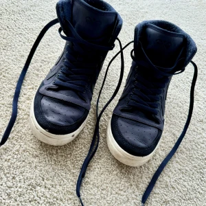 Mörkblå sko i Converse stuk med snörning - Säljer ett par Hummel, mörkblå sneakers, storlek 40, i skinn med snörning. De har en hög modell (converse). Sulan är vit och har en lite sliten look, vilket ger dem en cool, använd stil. Perfekta för både vardag och fest! Bra skick!