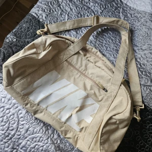 Beige axelväska från Nike - Säljer en snygg beige duffelba från Nike. Väskan har ett stort vitt Nike-tryck på sidan och är utrustad med en praktisk dragkedja. Perfekt för träning eller andra äventyr. Den är i syntetmaterial och har justerbara axelremmar för bästa passform!