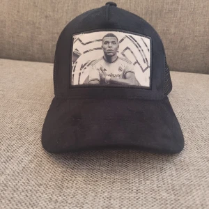 Svart keps från Evosportswear - Säljer en svart keps från Evosportswear med ett coolt tryck av en fotbollsspelare på framsidan. Kepsen har ett nätmaterial på baksidan för bra ventilation och en justerbar rem för perfekt passform. Perfekt för fotbollsfans eller som en stilren accessoar till vardagsoutfiten.