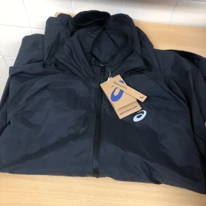 Svart vindjacka från Asics - Säljer en svart vindjacka från Asics med dragkedja och huva. Perfekt för utomhusaktiviteter med sin lätta och vattenavvisande design. Jackan har en enkel och stilren look med Asics-logga på bröstet.