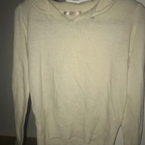 Beige tröja med pärldetaljer - Elegant beige tröja med pärlor längs kragen. Tröjan har långa ärmar och en mjuk, bekväm passform. Perfekt för en stilren look.