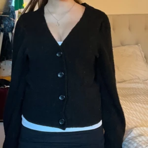 Svart kofta från & Other Stories - Säljer en stilren svart kofta från & Other Stories. Den har en v-ringad design med knappar framtill och långa ärmar. Perfekt för att hålla sig varm och snygg under kyligare dagar. Tröjan är nopprig men det är inget man tänker på.💕