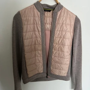 Grå och rosa jacka från Barbour - Snygg jacka från Barbour i en kombination av grått och rosa. Jackan har en quiltad framsida och rygg i rosa, med gråa ärmar och krage. Den är försedd med dragkedja och har en modern look som passar perfekt för vår. 