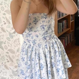 Blommig axelbandslös klänning - Charmig axelbandslös klänning med blått blommönster på vit bakgrund. Perfekt för sommarens varma dagar med sin luftiga design och korta längd.