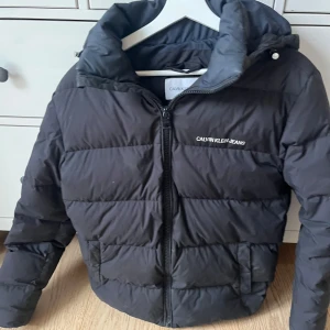 Svart dunjacka från Calvin Klein Jeans - Snygg svart dunjacka från Calvin Klein Jeans med dragkedja och huva. Jackan har en klassisk pufferdesign och logotypdetaljer på bröstet och ärmen. Perfekt för kyliga dagar. Jackan är använd men inte så illa fint skick 