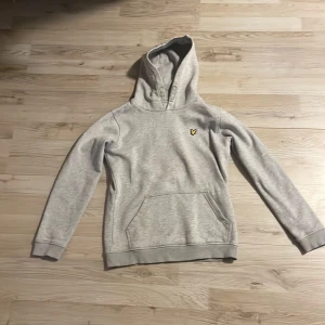 Grå hoodie med emblem - Säljer en stilren grå hoodie med ett litet emblem på bröstet. Tröjan har en klassisk känguruficka och en bekväm huva. Perfekt för en avslappnad look.