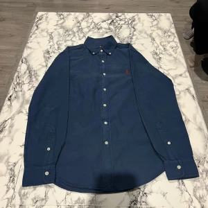 Blå skjorta från Ralph Lauren - Snygg blå skjorta från Ralph Lauren i slim fit. Skjortan har långa ärmar och knappar framtill. Den är tillverkad i bomull och har en röd broderad logotyp på bröstet.