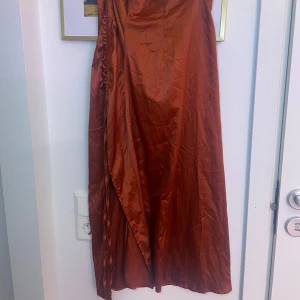 Rostfärgad långklänning - Elegant rostfärgad kjol i satin med omlott-design. Klänningen har en vacker glans och en smickrande passform. Perfekt för speciella tillfällen. Kjolen har slits på sidan med snören man kan dra åt! Kjolen har legat i en påse därav lite skrynklig