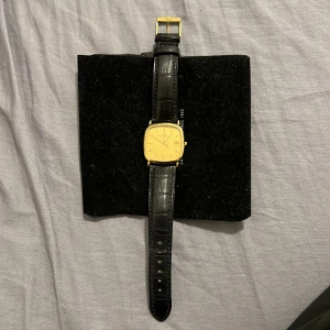 OMEGA Seamaster Gold Men's Watch - 1430 - Elegant guldfärgad klocka från Omega med fyrkantig urtavla och datumvisning. Klockan har ett svart läderarmband med krokodilpräglat mönster och ett guldfärgat spänne. Perfekt för den som söker en klassisk och stilren accessoar.