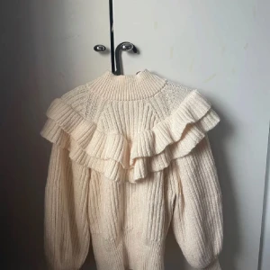 Beige stickad tröja med volang - Säljer en mysig beige stickad tröja med volangdetaljer runt axlarna. Tröjan har långa ärmar och en hög krage, perfekt för kyligare dagar. Den ribbade strukturen ger en fin textur och stil.