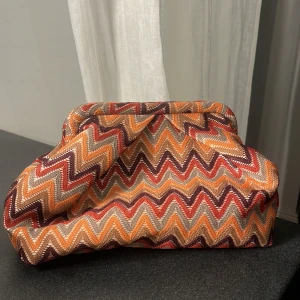 Mönstrad väska från Beck Söndergaard - Säljer en färgglad väska från Beck Söndergaard med ett zigzag-mönster i orange, röd och beige. Väskan har en dragkedja inuti och är perfekt för att hålla dina småsaker organiserade.