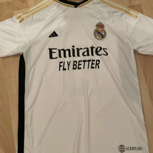 Vit Real Madrid fotbollströja - Säljer en vit Real Madrid fotbollströja från Adidas med korta ärmar och guldiga detaljer på axlarna. Tröjan har klubbens emblem och sponsorlogga på framsidan samt spelarnamnet Bellingham och nummer 5 på baksidan. Perfekt för fans av laget!