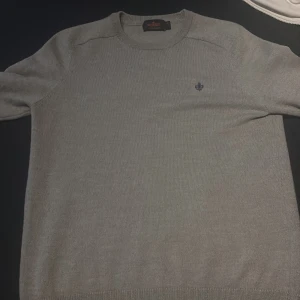 Grå tröja från Morris - Säljer en stilren grå tröja från Morris med rund hals och ett diskret broderat emblem på bröstet. Perfekt för en klassisk look.