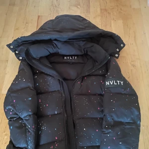Svart pufferjacka från NVLTY - Snygg svart pufferjacka från NVLTY med färgglada stänkdetaljer. Jackan har en huva och dragkedja framtill. Perfekt för kyliga dagar.